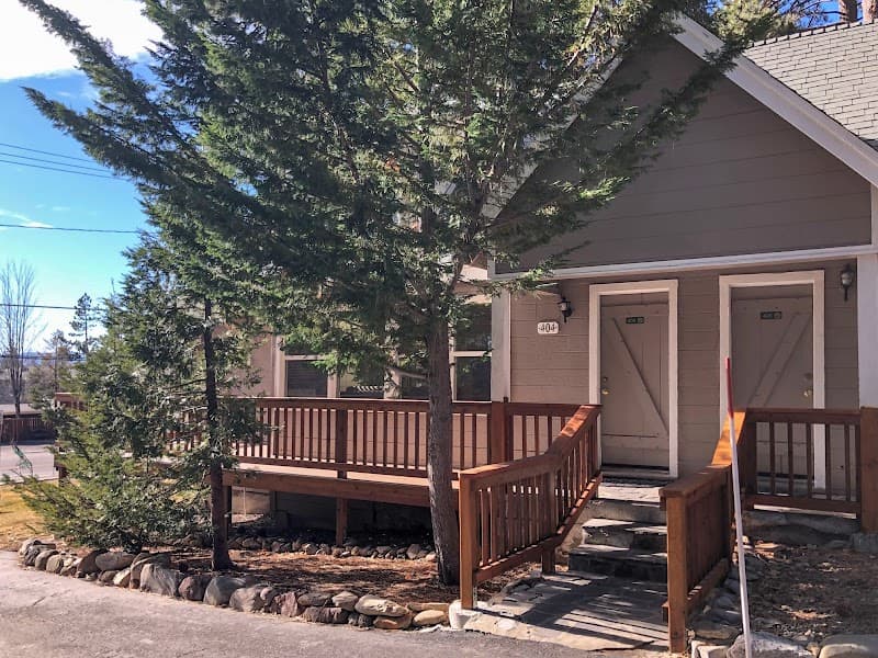 A serene lakeside resort scene features cozy cabins with wooden decks and stairs, surrounded by tall evergreen trees under a clear blue sky, ideal for tranquil family vacations.