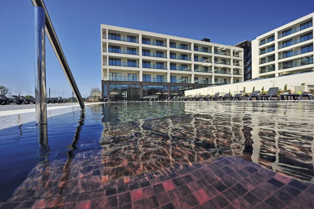 A serene hotel poolside scene with clear blue skies, modern multi-story buildings, lounge chairs, and reflective water, ideal for leisure travelers seeking relaxation.