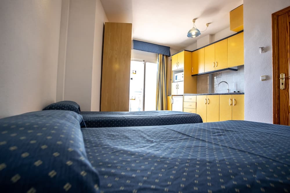 A cozy hotel room features blue patterned beds, a compact kitchenette with yellow cabinets, and a bright balcony view, ideal for travelers seeking comfort and convenience.