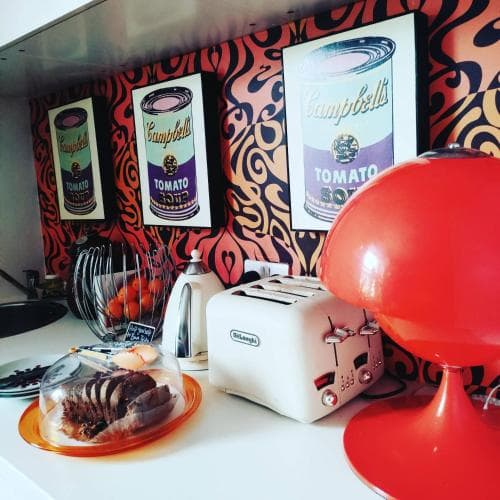 A vibrant retro kitchen scene featuring bold wallpaper, Warhol-inspired Campbell's Soup can prints, a sleek red lamp, and vintage appliances evokes a playful and artistic mood, perfect for travelers seeking unique and culturally rich accommodations.