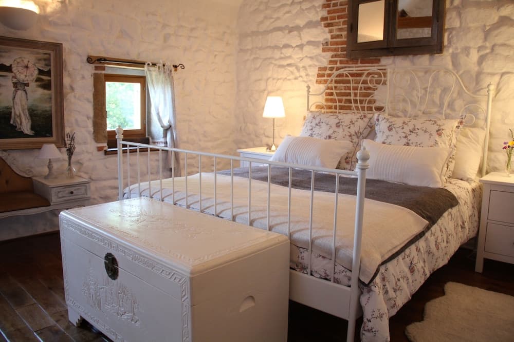 A cozy, rustic bedroom with stone walls, a white iron bed adorned with floral bedding, and a vintage trunk, ideal for travelers seeking a charming countryside retreat.