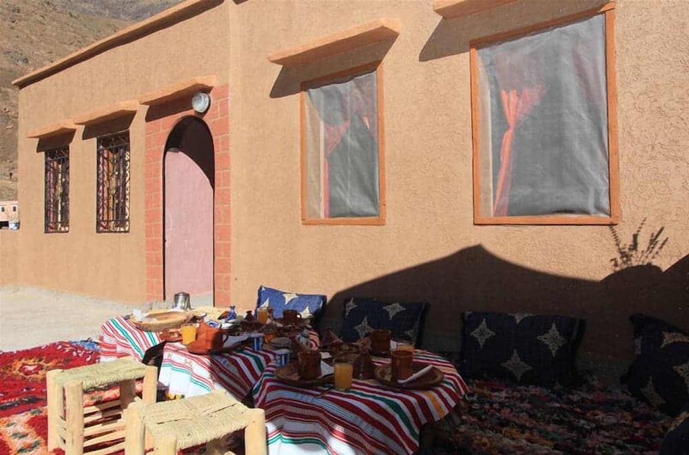 A cozy outdoor dining setup with colorful rugs, cushions, and traditional tableware invites travelers to relax in the serene ambiance of a rustic adobe house nestled against a mountainous backdrop, perfect for cultural immersion experiences.