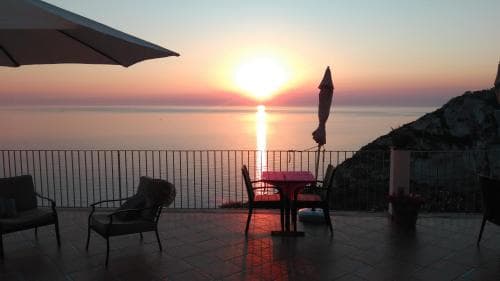 A serene sunset over the sea is captured from a cozy patio with lounge chairs and a table, ideal for travelers seeking relaxation and scenic views.