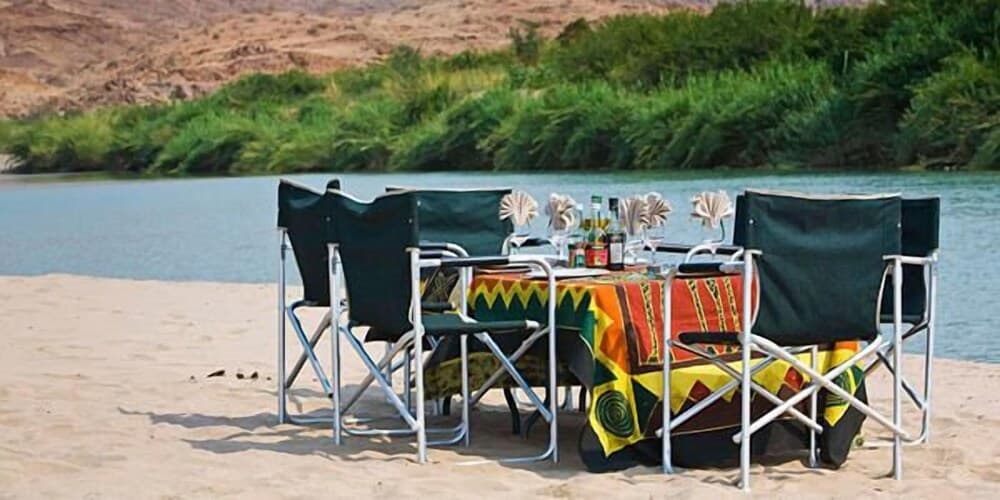 A serene riverside setting features a cozy outdoor dining area with folding chairs and a colorful tablecloth, perfect for a tranquil camping or picnic experience amidst lush greenery and sandy shores.