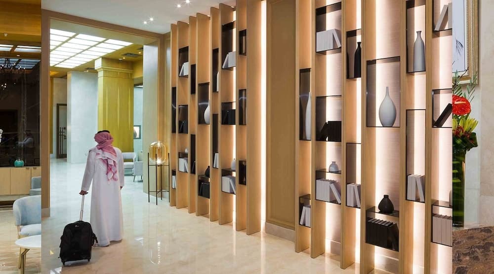 A traveler in traditional attire walks through a luxurious hotel lobby adorned with elegant wooden shelves displaying vases and art, creating a serene and sophisticated atmosphere ideal for business or leisure stays.