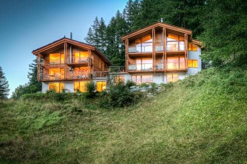 A cozy, warmly lit chalet nestled on a grassy hillside amidst tall pine trees offers a serene retreat for travelers seeking a peaceful mountain getaway.