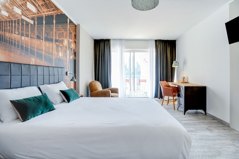 A cozy hotel room features a neatly made bed with green accents, a desk by the window with a view of balconies, and a wall mural, ideal for travelers seeking comfort and style.