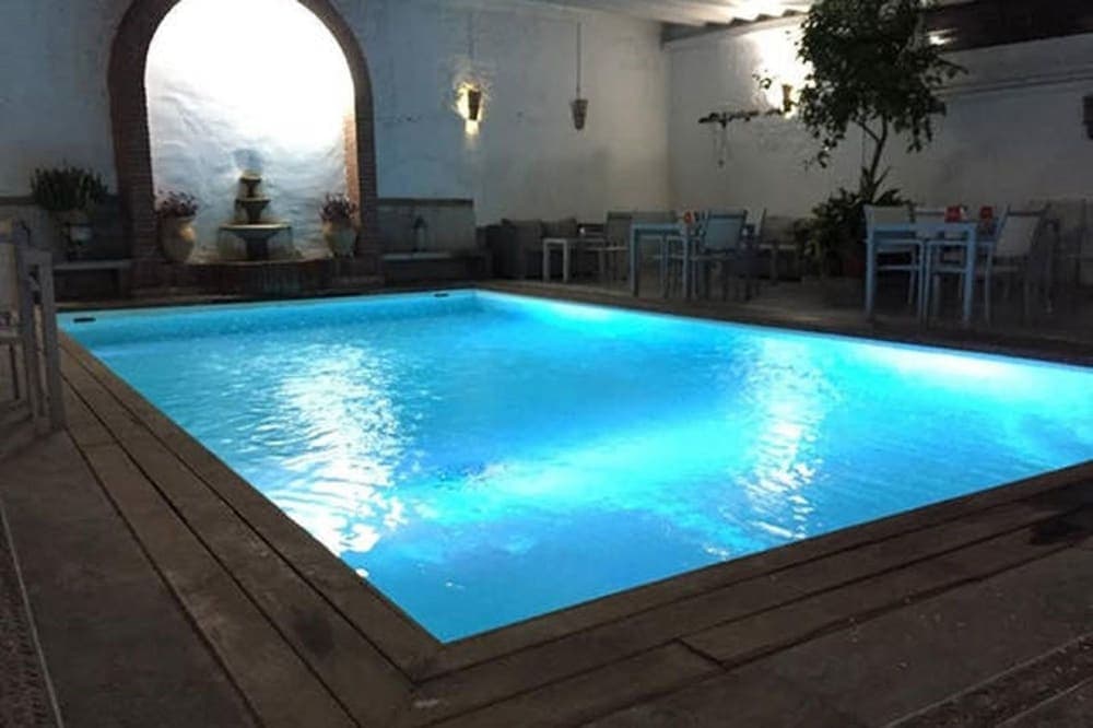 A serene indoor pool area illuminated by soft lighting features a glowing blue pool, cozy seating, and decorative elements like a fountain and plants, ideal for relaxation and leisure in a boutique hotel setting.