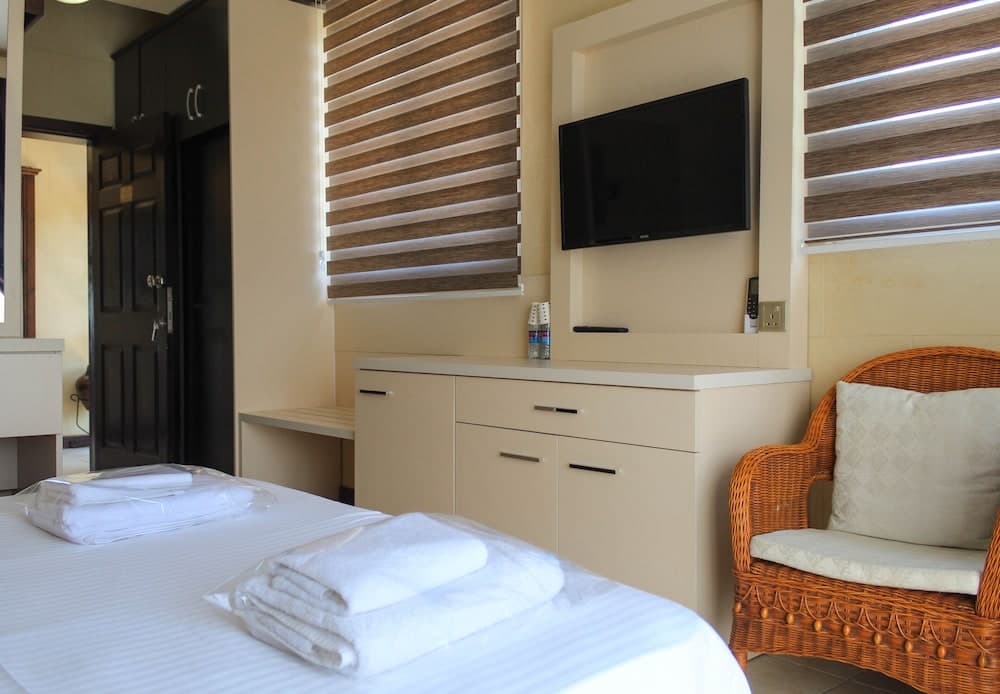 A cozy hotel room features a neatly made bed with folded towels, a wicker chair, a mounted TV, and wooden blinds, ideal for travelers seeking comfort and relaxation.