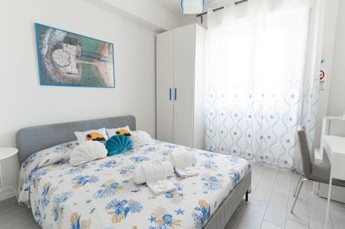 A cozy bedroom with a nautical-themed bedspread, soft lighting through sheer curtains, and a vibrant wall art piece creates a serene retreat ideal for travelers seeking a relaxing seaside accommodation.