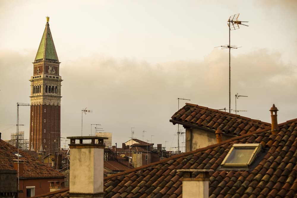 A serene rooftop view of Venice showcases tiled roofs, chimneys, antennas, and the iconic bell tower under a soft, cloudy sky, ideal for travelers seeking historical ambiance and panoramic vistas.