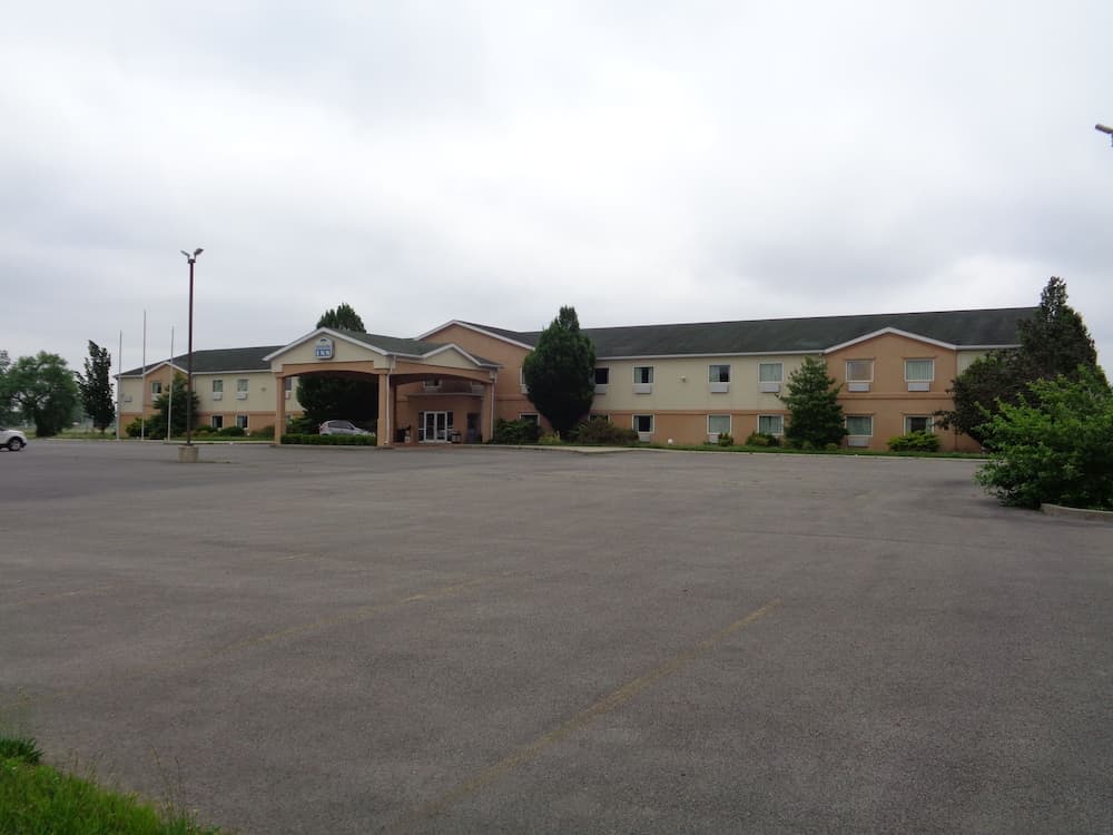A quiet motel with a spacious parking lot under an overcast sky, featuring a covered entrance and lush greenery, ideal for travelers seeking a simple accommodation stop.
