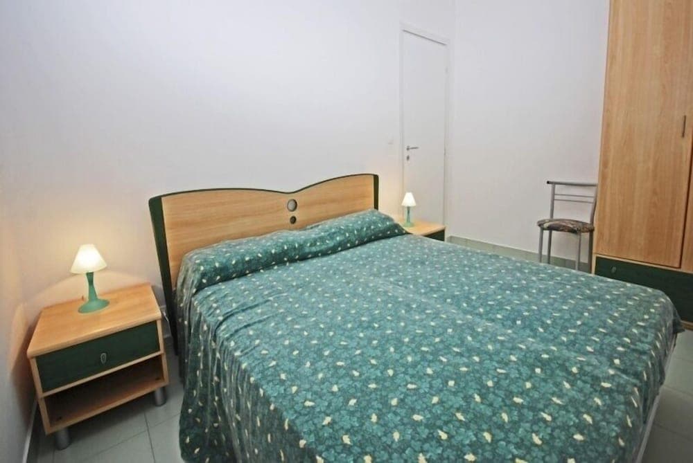 A cozy bedroom with a neatly made bed featuring a green patterned duvet, wooden furniture including bedside tables with lamps, and a calm ambiance ideal for travelers seeking a simple and comfortable stay.