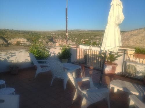 A serene rooftop terrace with white chairs, potted plants, and a closed umbrella overlooks a picturesque valley under a clear blue sky, ideal for travelers seeking relaxation and scenic views.