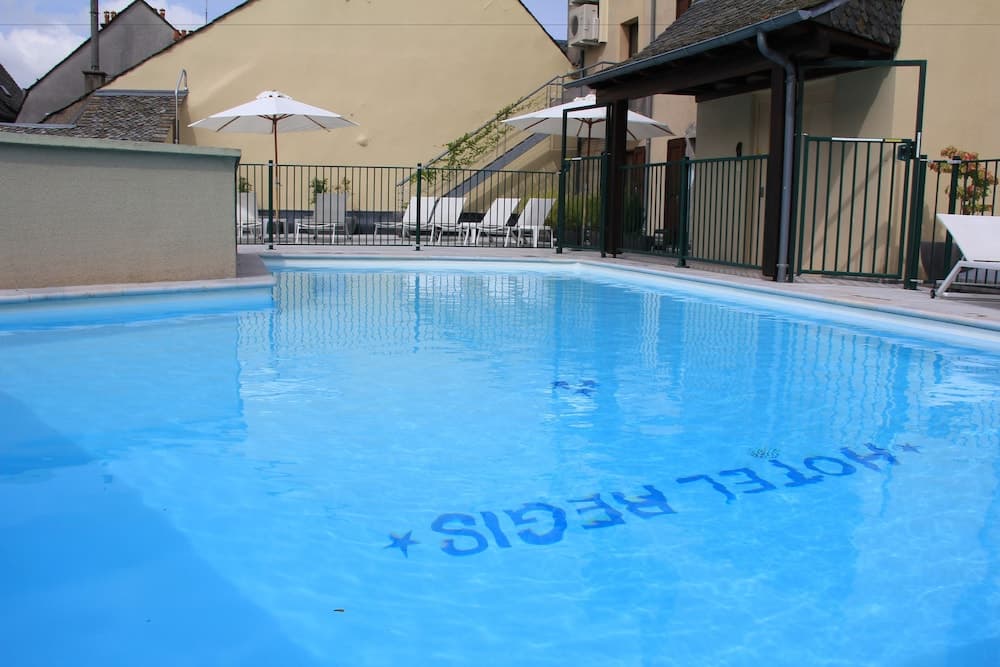 A serene outdoor swimming pool with "HOTEL REGIS" written on the bottom, surrounded by lounge chairs, umbrellas, and a fenced patio area, ideal for travelers seeking relaxation in a cozy hotel setting.