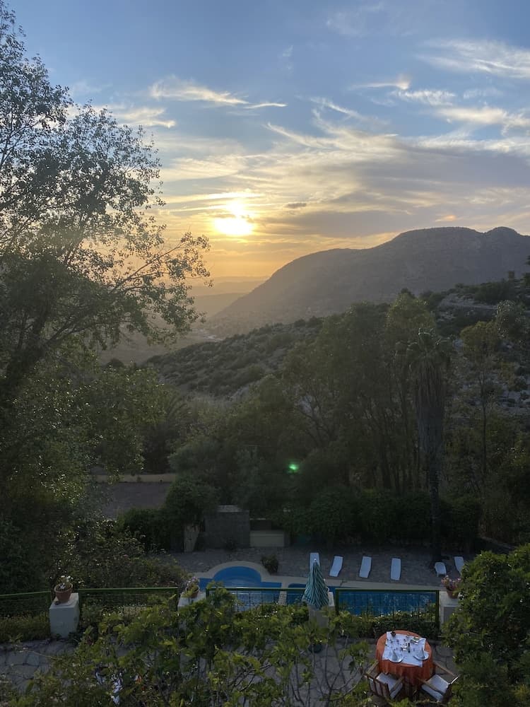 A serene sunset over a lush hillside frames a tranquil poolside area with lounge chairs and greenery, ideal for a relaxing vacation retreat.
