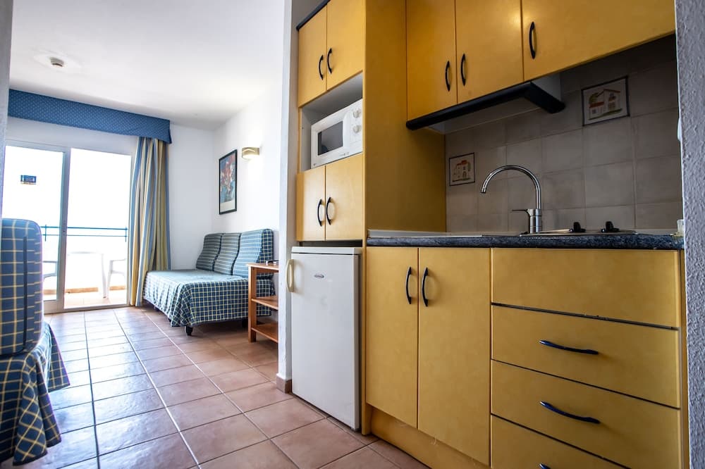 A cozy apartment kitchenette with yellow cabinets, a compact fridge, and a view of a tiled living area leading to a balcony, ideal for travelers seeking self-catering accommodations.