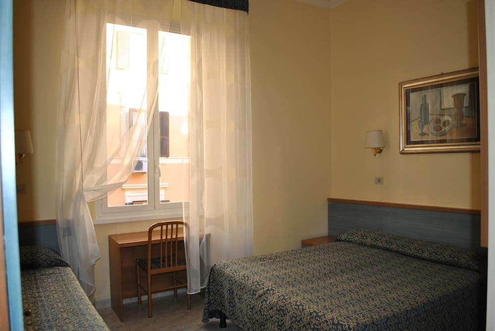 A cozy hotel room with soft yellow walls, patterned bedding, sheer curtains framing a bright window, and a small desk by the bed, ideal for travelers seeking comfort and simplicity.