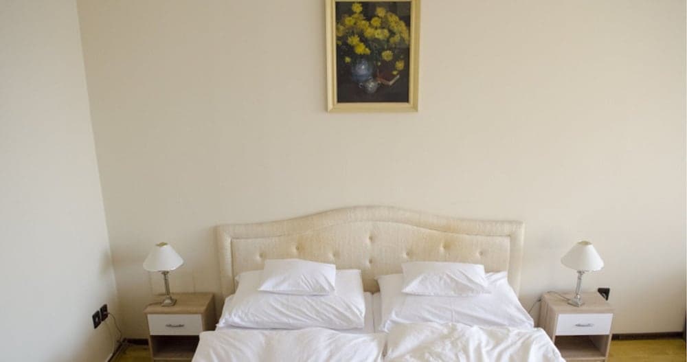 A cozy hotel bedroom features a neatly made double bed with white linens, flanked by matching bedside tables with lamps, under a floral painting, ideal for travelers seeking comfort and simplicity.