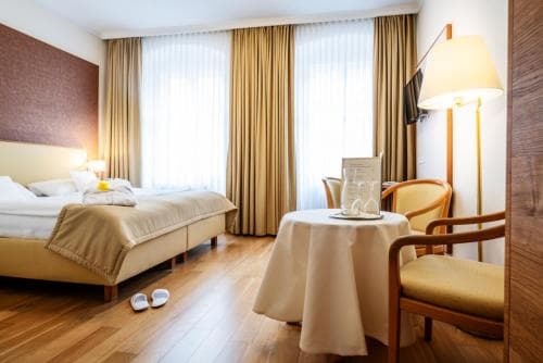 A cozy hotel room features a neatly made bed, wooden flooring, a small table with glasses, and warm lighting, creating an inviting atmosphere ideal for travelers seeking comfort and relaxation.