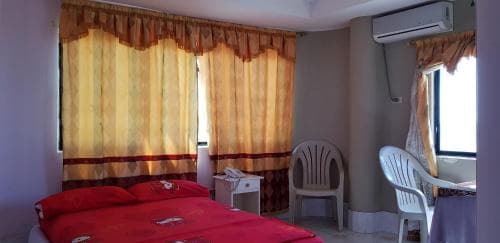 A cozy hotel room features a neatly made bed with red bedding, two plastic chairs, a small table, patterned curtains, and an air conditioner, ideal for travelers seeking a comfortable and budget-friendly stay.