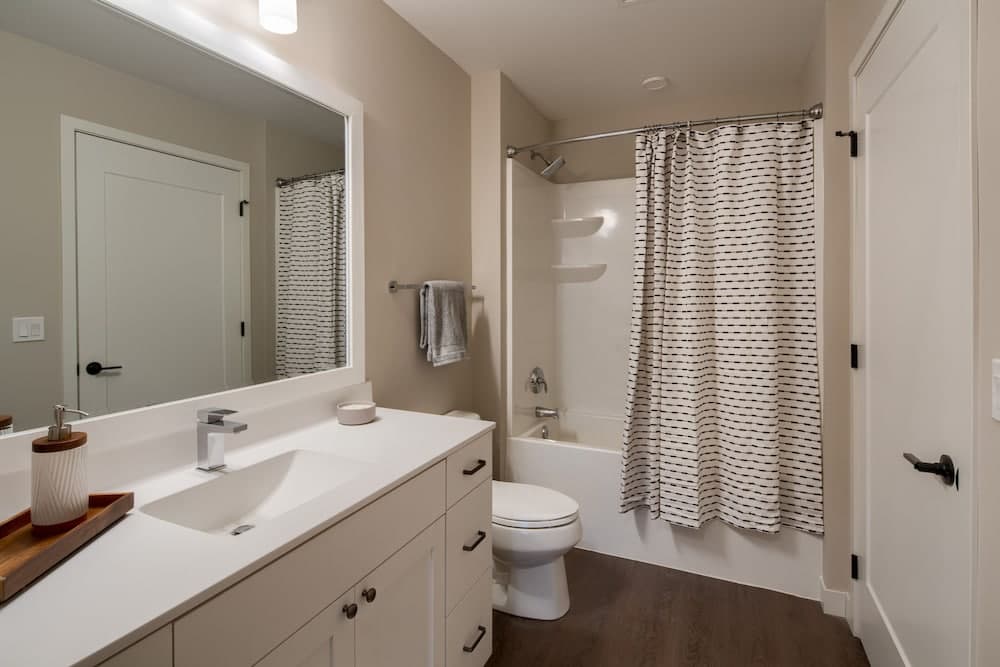 A cozy bathroom with beige walls, a white vanity, striped shower curtain, and wooden flooring creates a serene atmosphere ideal for travelers seeking comfort and cleanliness in their accommodations.