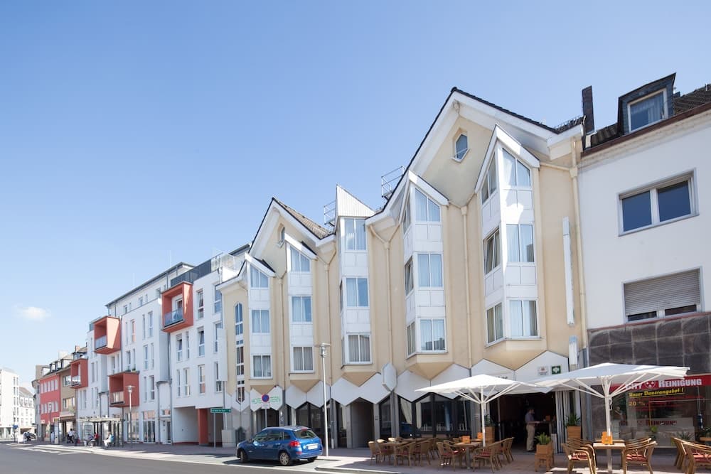 A vibrant European street scene features modern, colorful apartment buildings under a clear blue sky, with outdoor café seating and a parked car, ideal for urban exploration and leisurely dining.