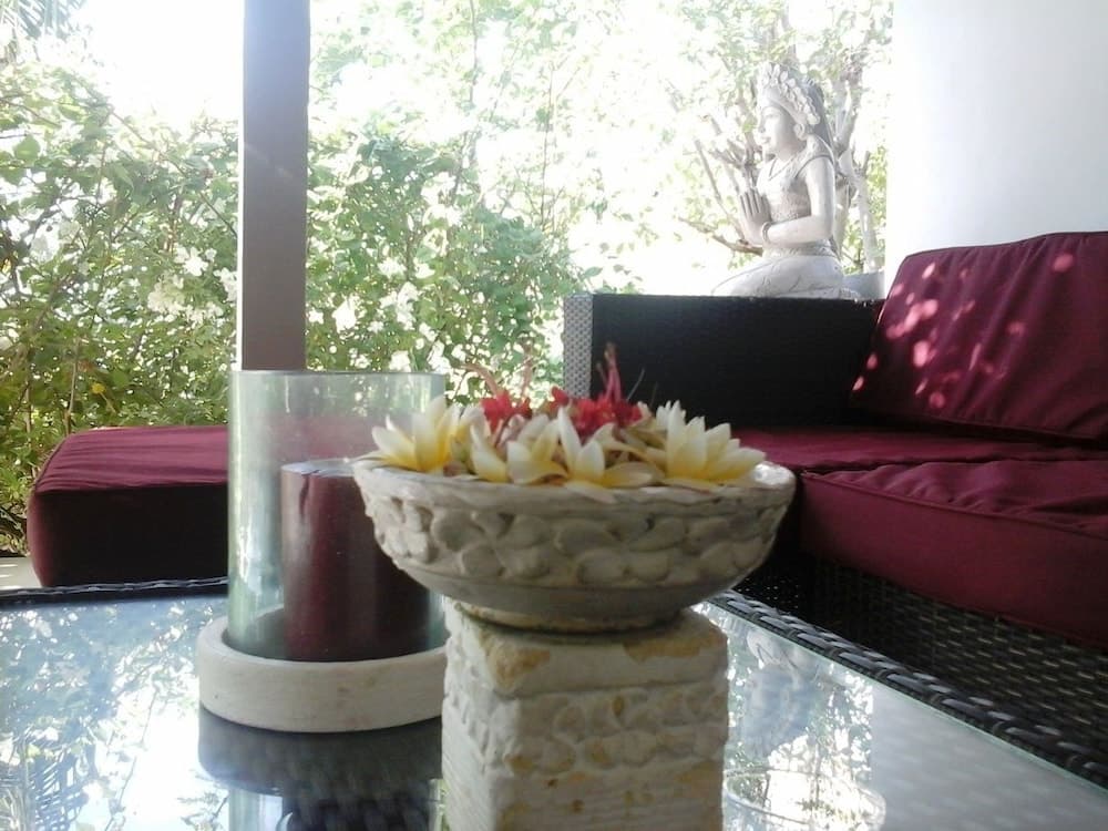A serene outdoor patio features a glass table adorned with a bowl of white flowers and a candle, set against lush greenery and a meditative statue, ideal for relaxation and spiritual retreats.