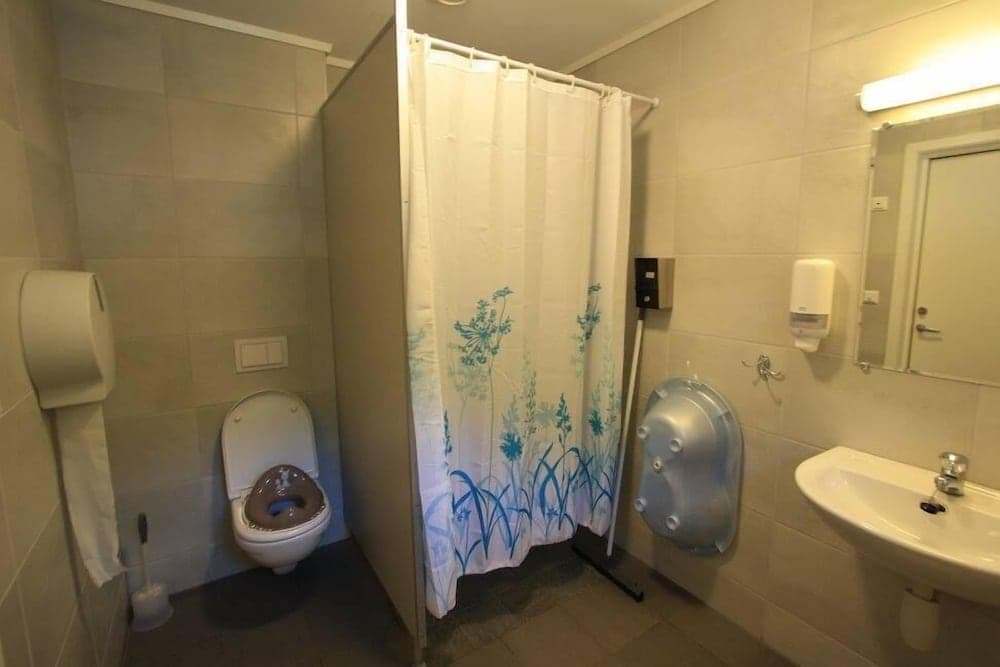 A compact bathroom with beige tiled walls features a toilet, sink, shower with a floral curtain, and a wall-mounted soap dispenser, ideal for travelers seeking basic amenities in a functional space.