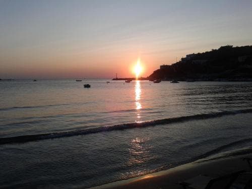 A serene sunset over a coastal beach with gentle waves, silhouetted boats, and a hillside town in the background creates a tranquil atmosphere ideal for relaxation and photography.