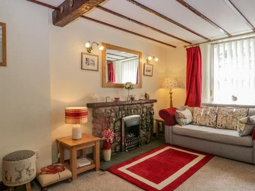 A cozy living room with exposed wooden beams, a stone fireplace, red accents, and warm lighting creates an inviting atmosphere ideal for a relaxing getaway in a countryside cottage.