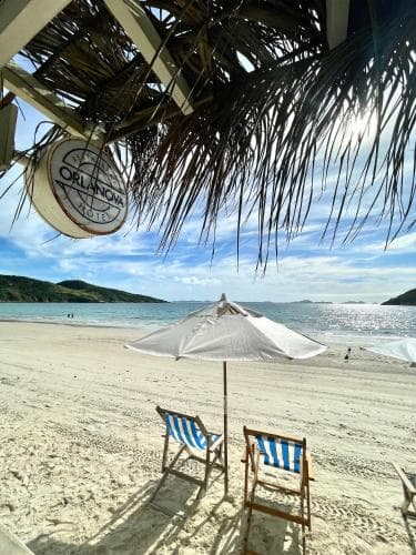 A serene tropical beach scene features two striped lounge chairs under a white umbrella, framed by palm fronds and a clear blue sky, ideal for a relaxing vacation getaway.