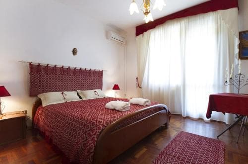 A cozy hotel room features a neatly made bed with red patterned bedding, soft lighting from a chandelier, and a warm ambiance ideal for travelers seeking comfort and relaxation.