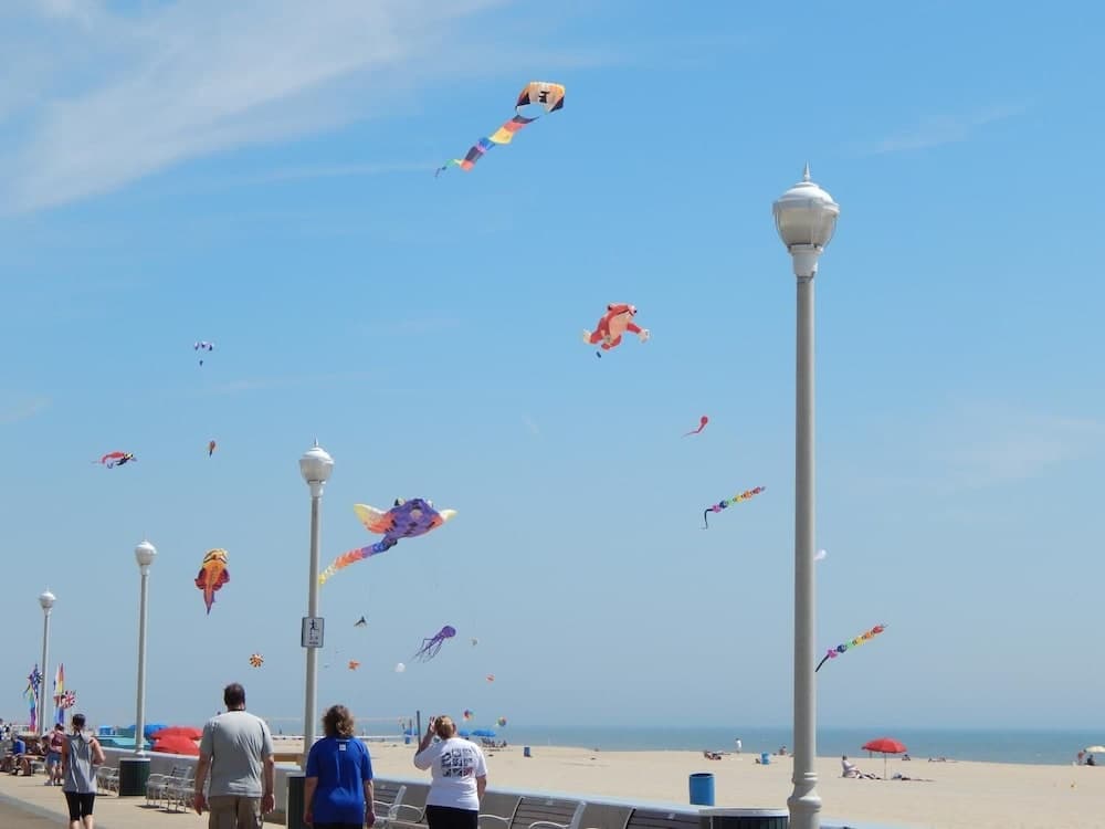 A vibrant beach scene with colorful kites soaring in the clear blue sky, lampposts lining the boardwalk, and visitors enjoying the sunny day, perfect for family outings and kite-flying enthusiasts.