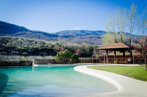 A serene outdoor pool area surrounded by lush greenery and distant mountains under a clear blue sky, featuring a wooden gazebo and blooming trees, ideal for a tranquil vacation retreat.
