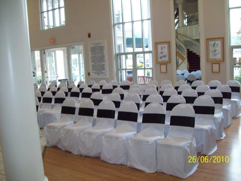 A spacious event hall with rows of white chairs adorned with black sashes, large windows letting in natural light, framed artwork on the walls, and a staircase in the background, ideal for wedding ceremonies or formal gatherings.