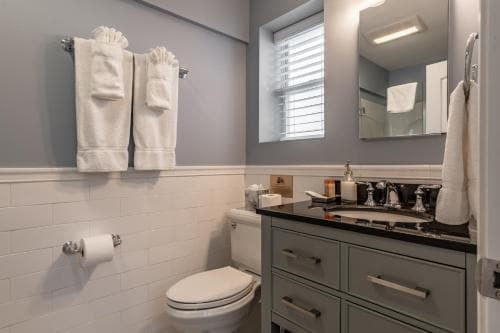 A cozy bathroom with soft gray walls, white subway tiles, neatly folded towels on a towel rack, and a sleek vanity with a black countertop, ideal for travelers seeking comfort and cleanliness in their accommodations.