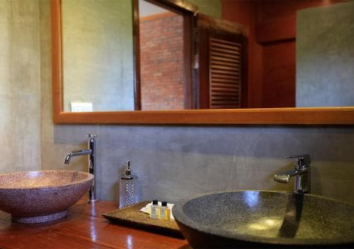 A serene bathroom with rustic stone sinks, a wooden vanity, and a large mirror reflecting brick walls, ideal for travelers seeking a tranquil and eco-friendly accommodation experience.