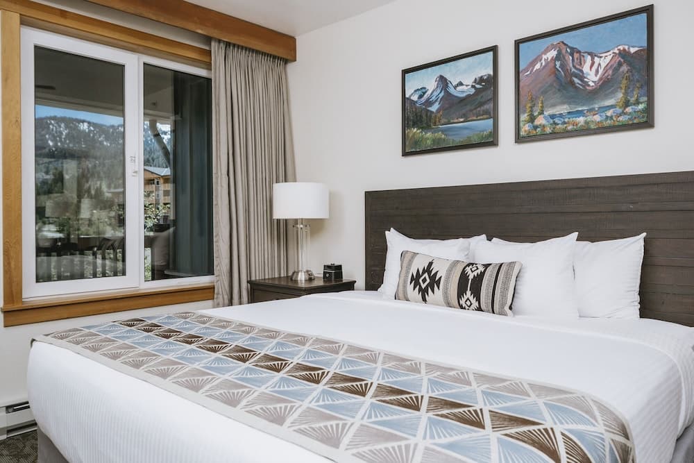 A cozy hotel room with a neatly made bed adorned with patterned linens, framed mountain artwork on the walls, and a serene view of snowy peaks through large windows, ideal for travelers seeking relaxation in a picturesque mountain retreat.