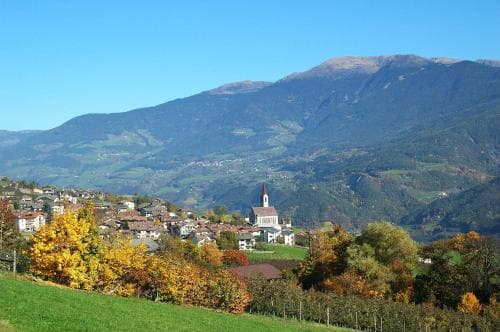 A picturesque village nestled among rolling hills and autumn-colored trees under a clear blue sky, featuring a prominent church spire, ideal for scenic photography and tranquil mountain retreats.