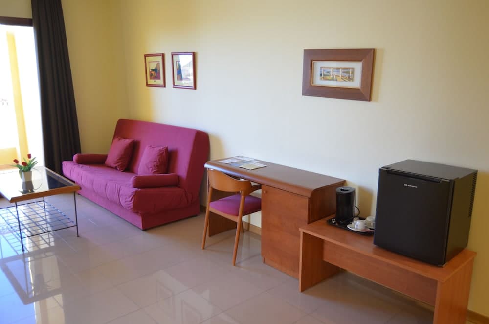 A cozy hotel room features a vibrant red sofa, wooden desk with a coffee maker, mini fridge, and framed artwork on the walls, creating a welcoming atmosphere ideal for travelers seeking comfort and convenience.