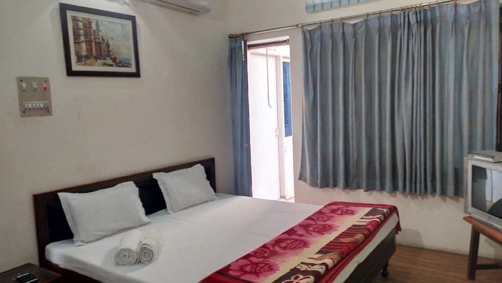 A cozy hotel room with a neatly made bed adorned with floral bedding, blue curtains framing a bright doorway, and a framed painting on the wall, ideal for travelers seeking comfort and simplicity.