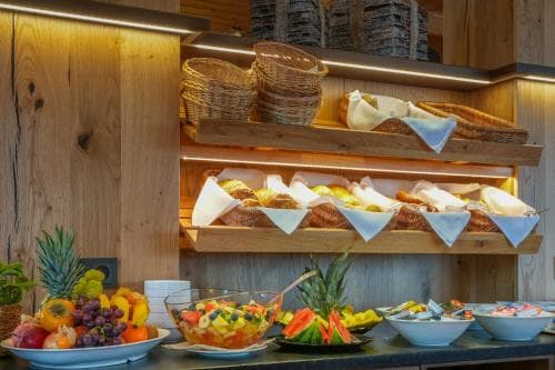 A cozy breakfast buffet area with wooden shelves displaying assorted breads and pastries under warm lighting, accompanied by vibrant fruit platters and tropical decor, evokes a welcoming morning ambiance ideal for travelers seeking a refreshing start to their day.