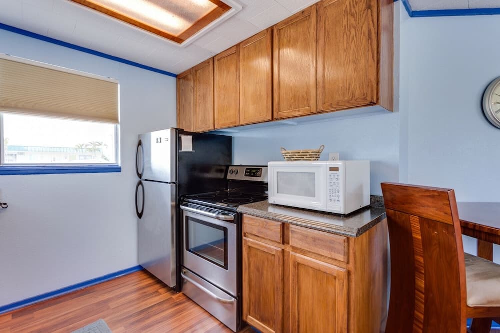 A cozy kitchenette with wooden cabinets, stainless steel appliances, and a window view of palm trees offers a comfortable space for travelers seeking a homey retreat.