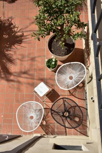 A cozy outdoor patio features stylish wicker chairs and tables surrounded by lush potted plants on a brick floor, creating an inviting and serene atmosphere perfect for relaxation or intimate gatherings.