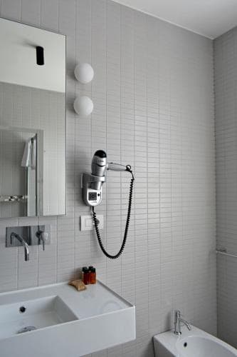 A modern, minimalist bathroom with white tiled walls, a sleek sink, a mounted hairdryer, and spherical wall lights creates a clean and serene atmosphere, ideal for travelers seeking a refreshing stay.