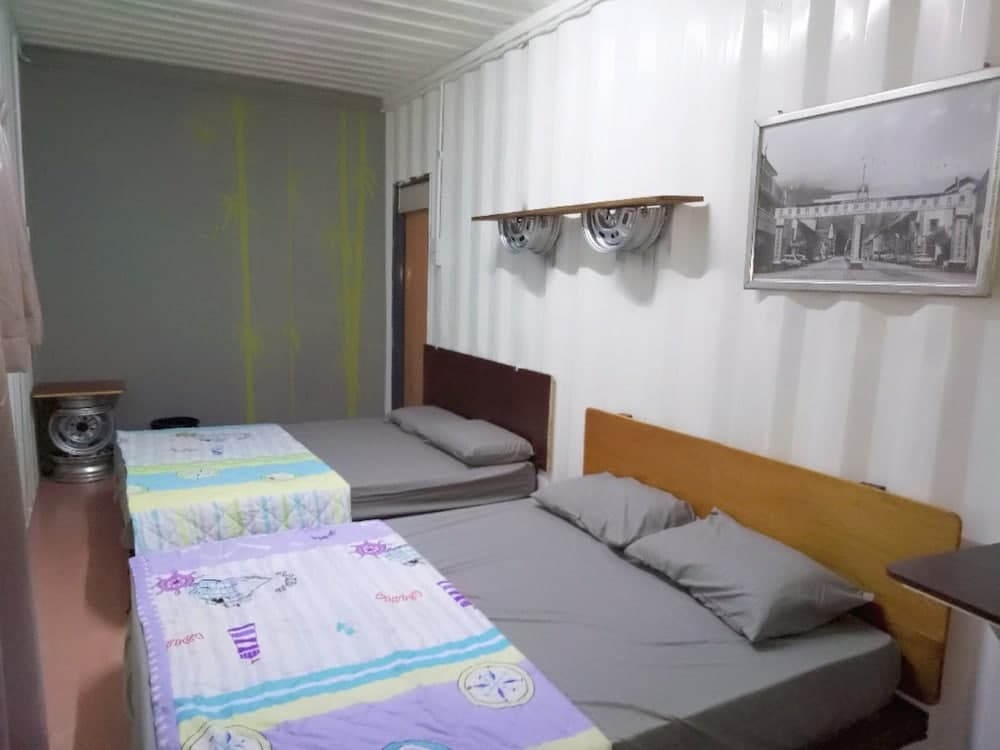 A cozy shipping container bedroom features two neatly made beds with colorful bedding, a vintage-style heater, and a framed black-and-white photograph, ideal for budget travelers seeking unique accommodations.