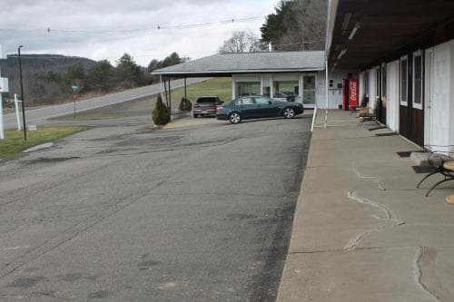 A quiet roadside motel with a covered parking area, cars parked nearby, and a serene mountain backdrop offers travelers a peaceful stopover in a rural setting.