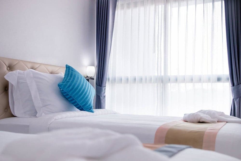 A serene hotel room features a neatly made bed with white linens and a blue pillow, soft natural light filtering through sheer curtains, ideal for travelers seeking comfort and relaxation.