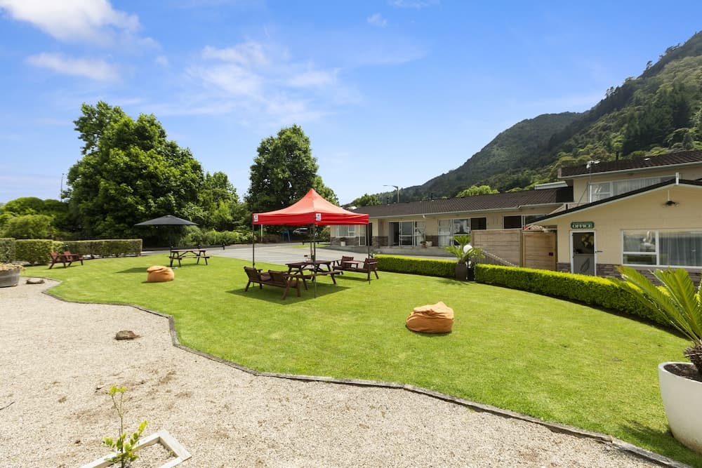 A serene outdoor recreational area features a vibrant green lawn with picnic tables under a red canopy, surrounded by lush trees and a gravel pathway, ideal for family-friendly vacations near mountainous landscapes.